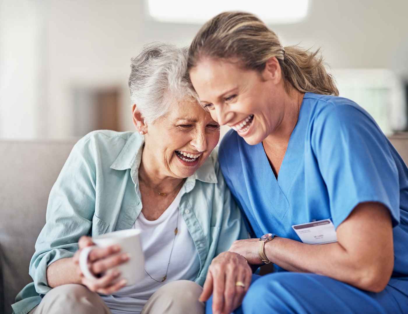 Warm companion PCA sharing joyful conversation and support with smiling senior woman on couch at home
